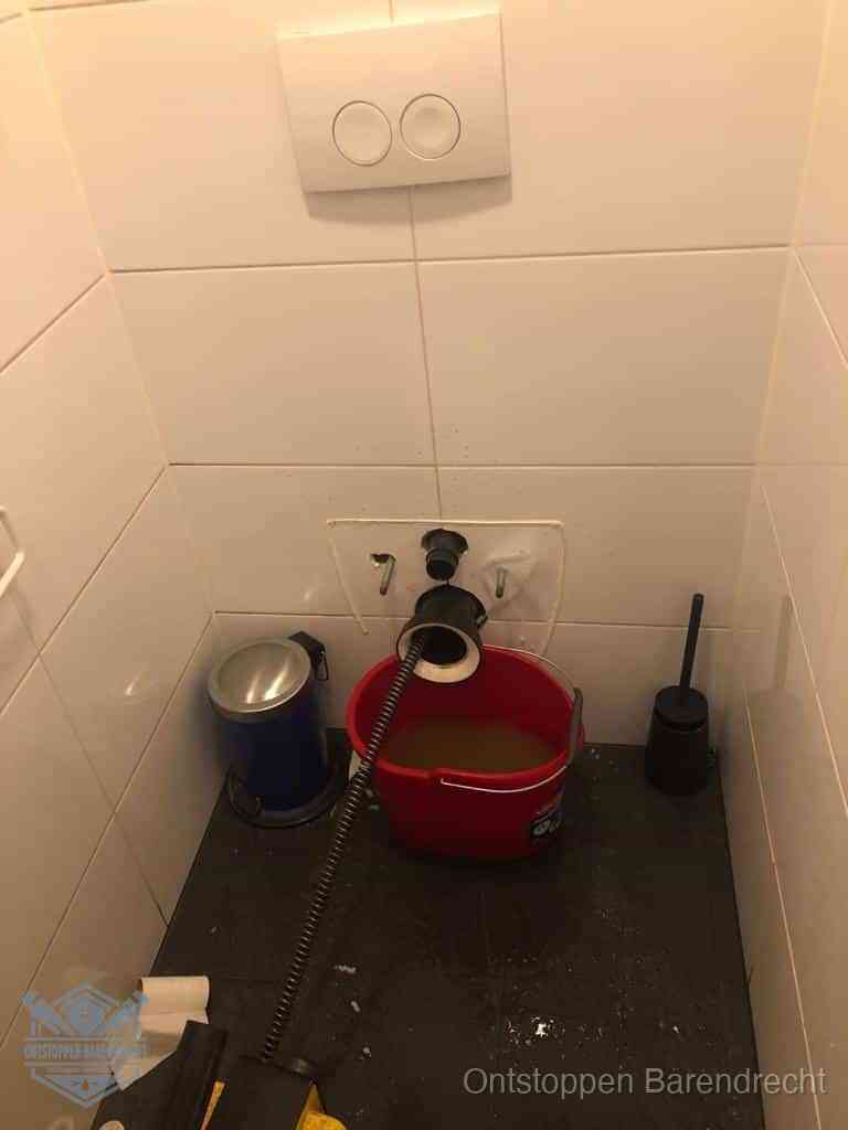 Een ontstopte wand-wc met verwijderde toiletpot waar een ontstoppingsveer in de wanduitgang verdwijnt, het achterhalen waar de verstopping zit in Barendrecht