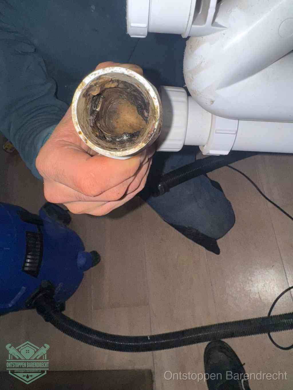 Een hand houdt een losgekoppeld PVC-afvoerbuisstuk vast dat van binnen volledig is dichtgeslibd met vet, vuil en aanslag, de meest voorkomende oorzaak van een verstopte afvoer in Barendrecht