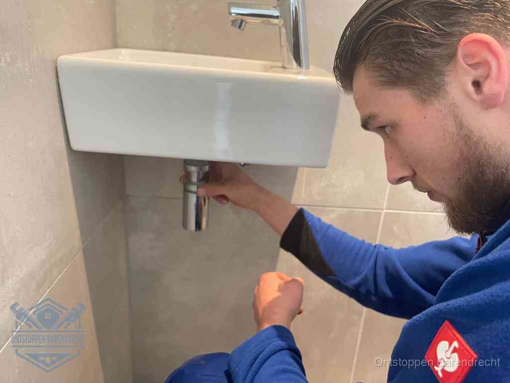 Een monteur draait met de hand de zwanenhalssifon onder een fonteinwasbak in de toiletruimte los om de bron van rioollucht in Barendrecht te vinden