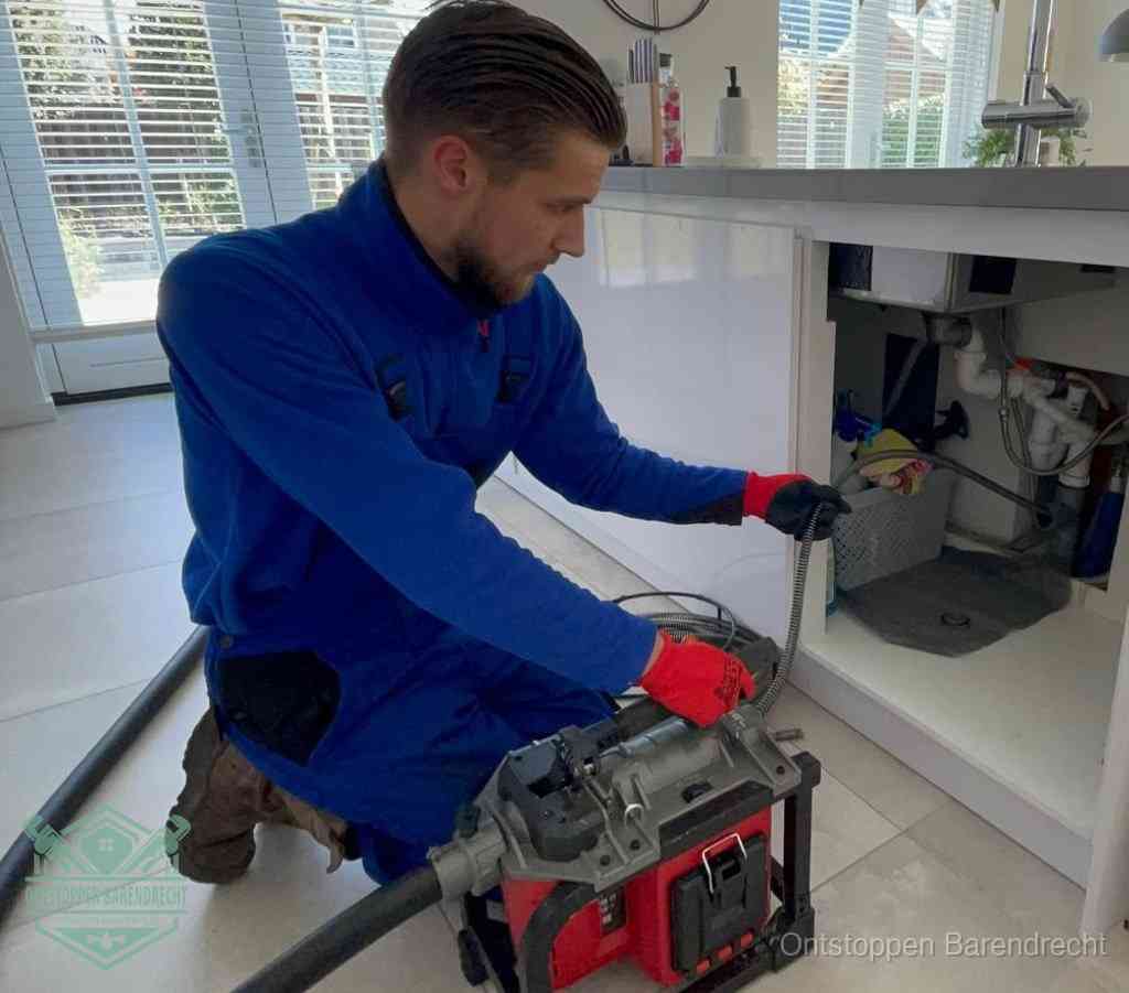Een monteur met rode handschoenen knielt in een keuken en voert een elektrische ontstoppingsveer in tijdens het professioneel afvoer ontstoppen in Barendrecht