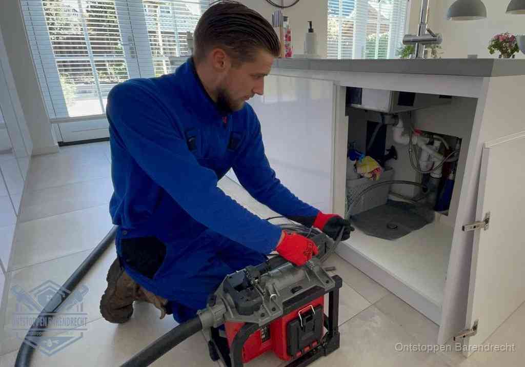 Een monteur voert een professionele elektrische ontstoppingsmachine in de afvoer onder de spoelbak tijdens het gootsteen ontstoppen in Barendrecht